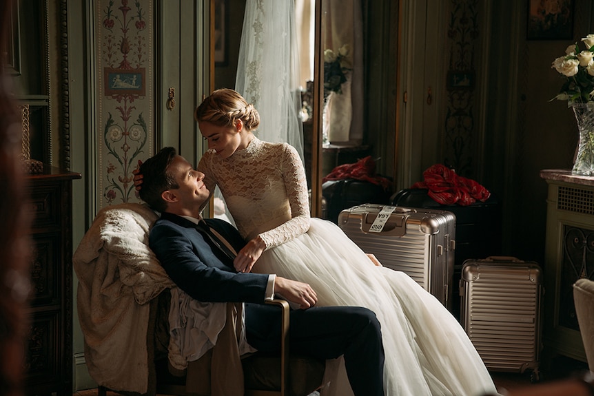 Samara Weaving in lace wedding dress sits on the lap of Mark O'Brien in suit, both look lovingly towards each other in room.