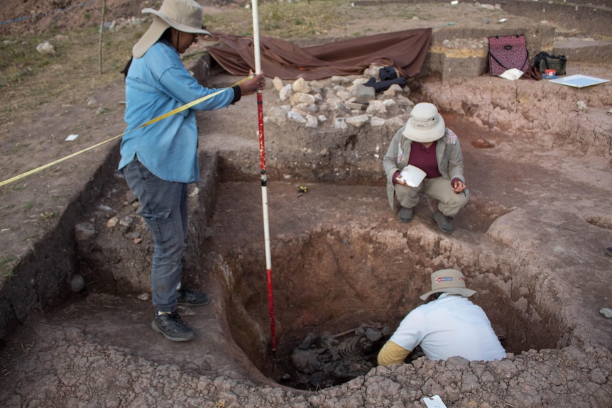 Three people gathered around a hole in the ground one standing holding a stick one kneeling and the third inside