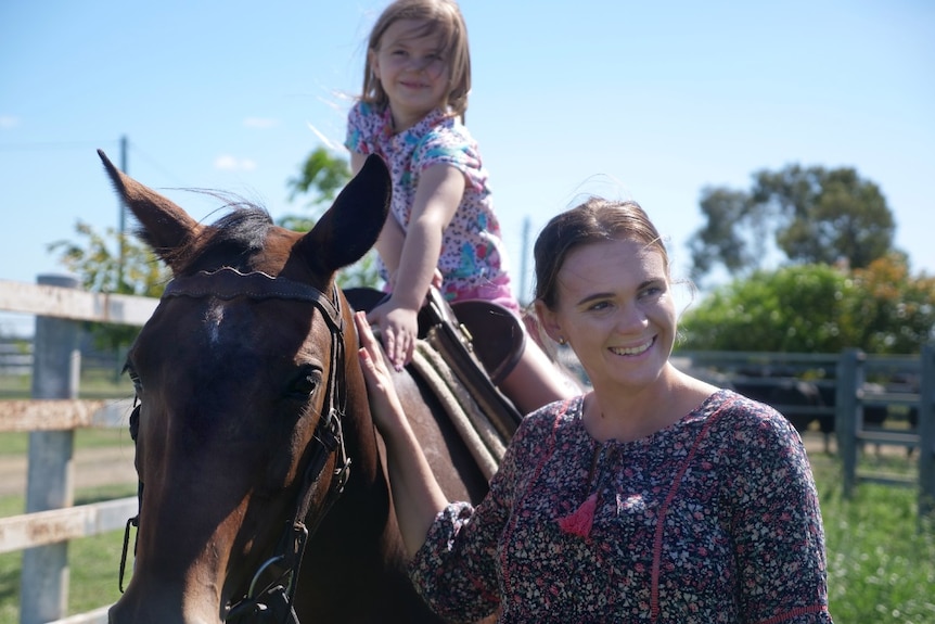 Samantha Comiskey standing, hand on horse, smiling, daughter sitting on horse.