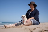 A middle-aged woman sitting on the beach gazing out to sea with a small dog sitting in front of her.