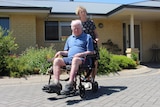 Norman sits in a wheelchair and Elaine stands behind him on a sunny day. They are not smiling. 