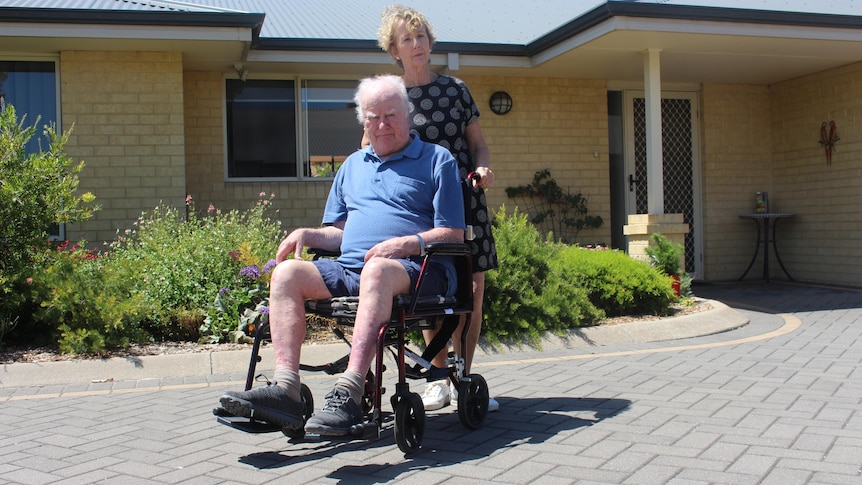 Norman sits in a wheelchair and Elaine stands behind him on a sunny day. They are not smiling. 