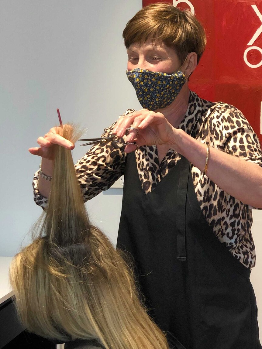 A woman in a mask standing in front of a red artwork cuts a woman's hair.