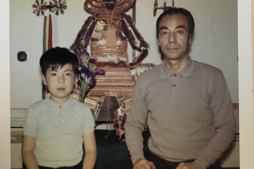 A young boy and man kneel on cushions in front of a small shrine.