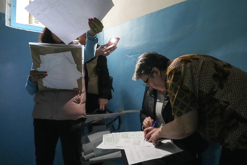 A woman signs a ballot paper as other sort through papers in a room.