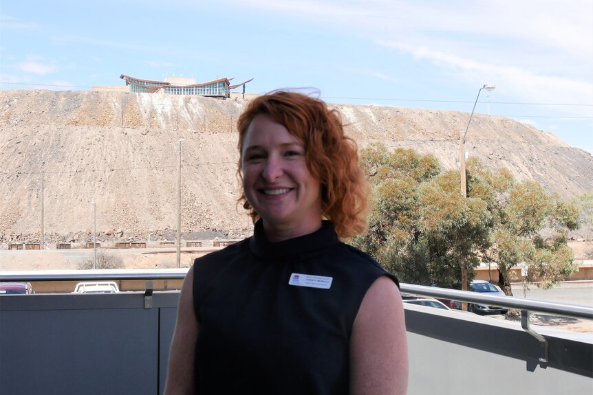 A woman with short red hair smiling, standing in front of large dirt mound in Broken Hill