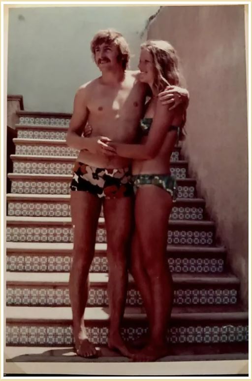 A young man and woman smile standing near steps in swimwear as they look away from camera.