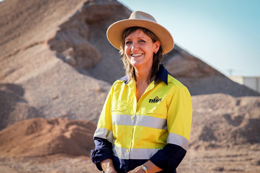 A woman in a brimmed hat and high vis standing in a quarry and smiling, in front of a pile of concrete.