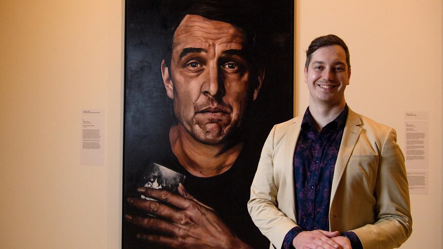 A man in a beige suit smiles next to a large painting of actor Samuel Johnson holding a black and white photo