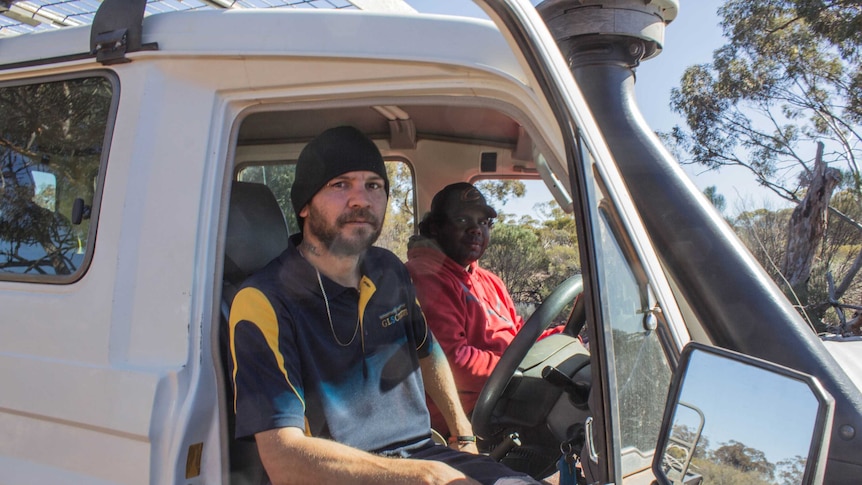 Two men sit in the front seat of a four wheel drive parked in bushland.