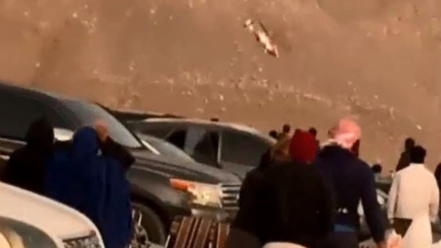 A group of around 10 bystanders watch next to three parked cars as a helicopter spirals before crashing on a mountain.