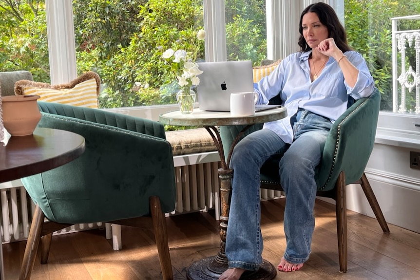 Erica Packer seated in a well-lit room with large glass windows. She sits at a laptop
