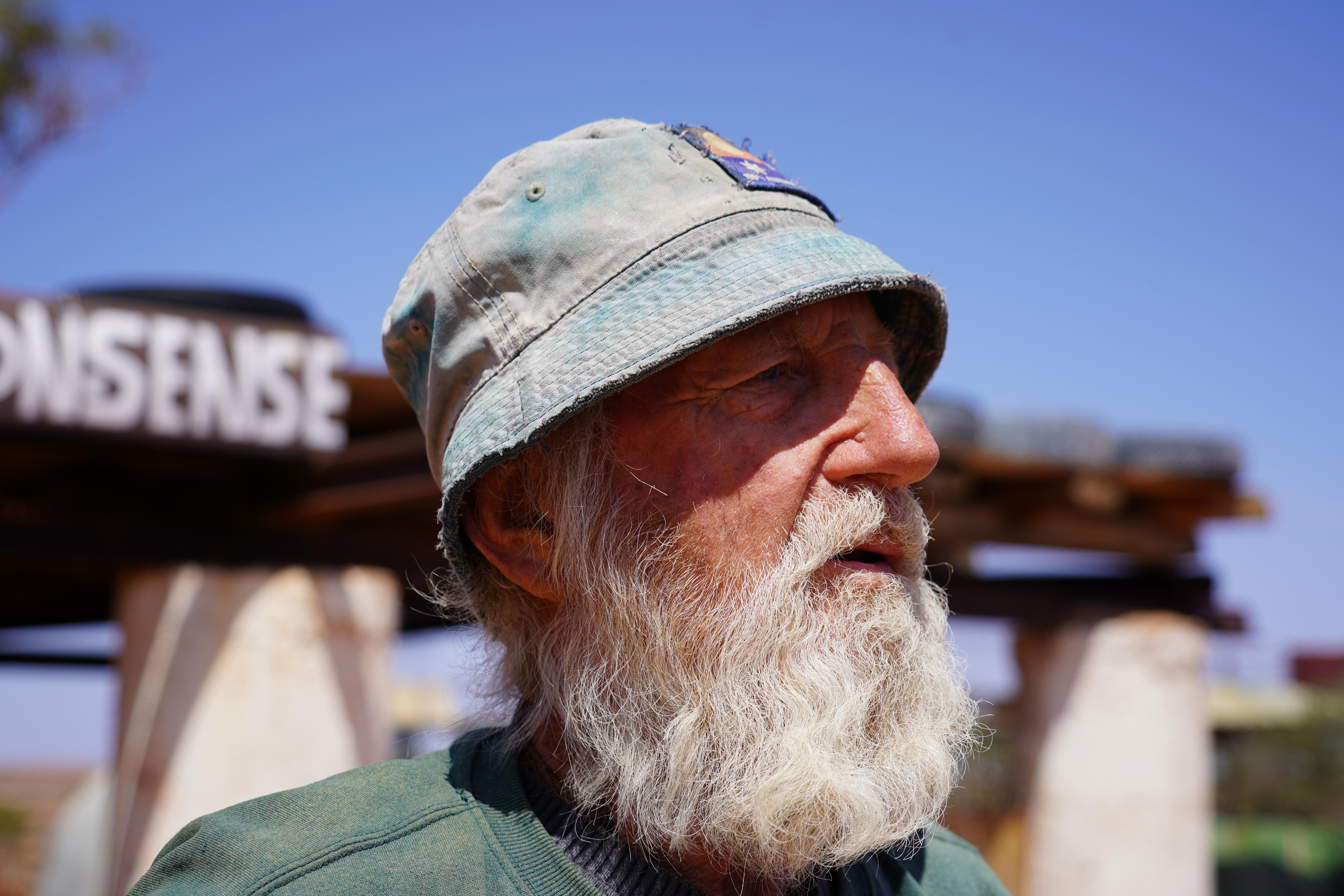 Un primer plano del rostro de un hombre mirando hacia un lado. Tiene una larga barba blanca y usa un sombrero de pescador. 