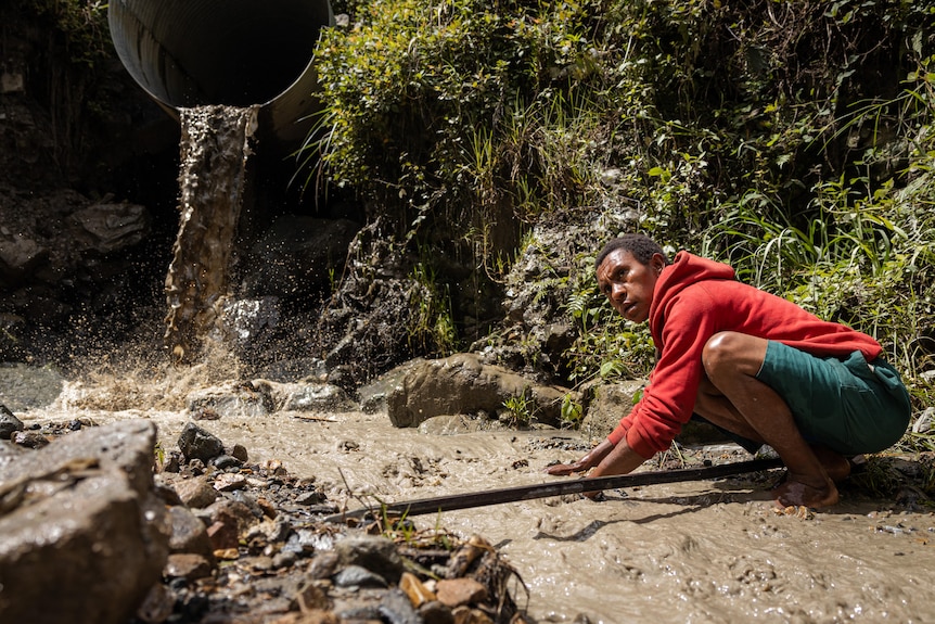 A person in a red hoodie crouches next to a drain flowing into a waterway 