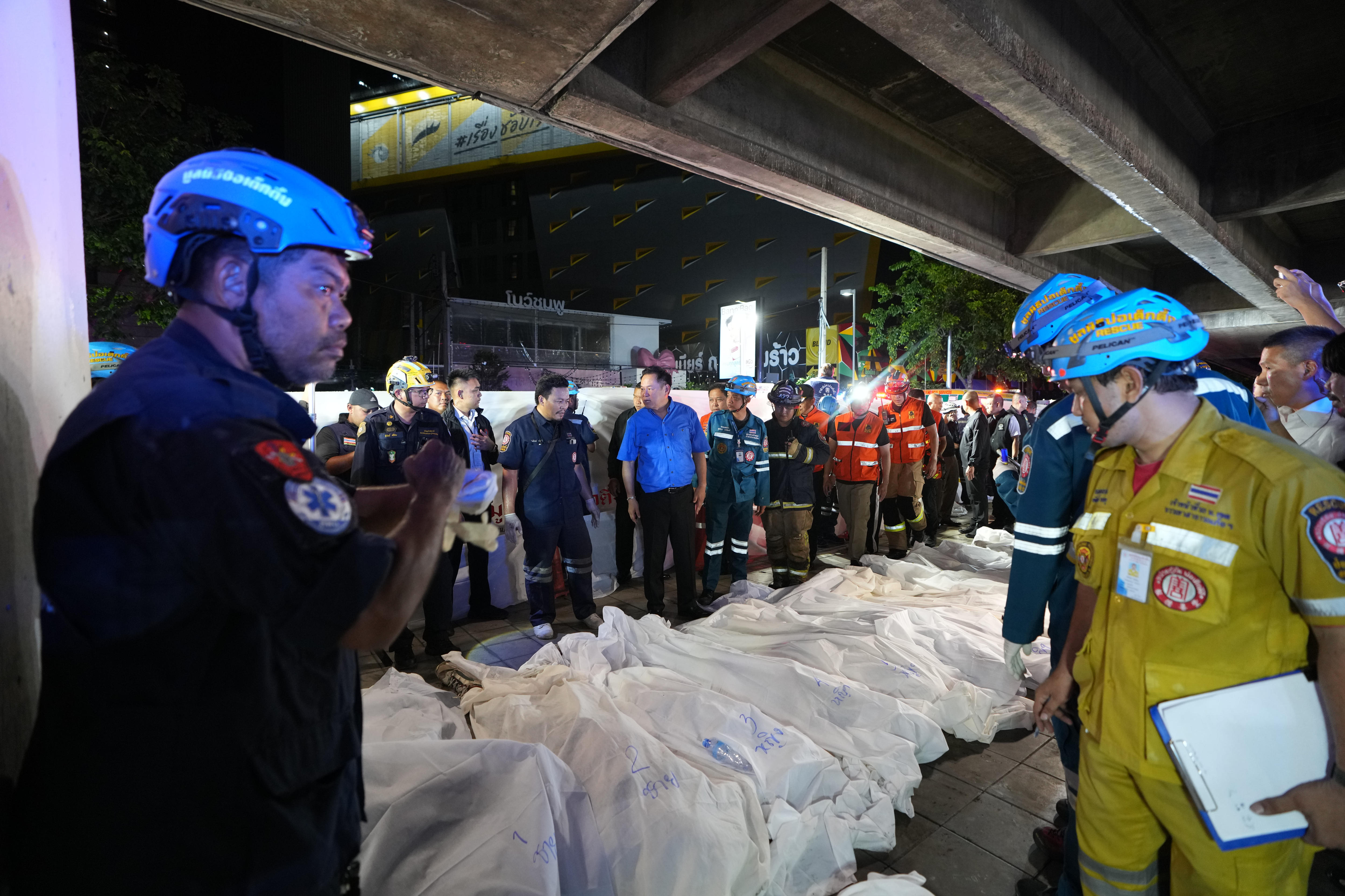 A row of bodies, covered in sheets, lie on the ground as first responders stand around.