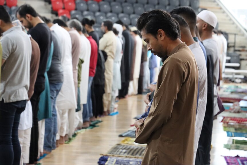 A large group of people with their heads bowed in prayer, standing up, inside an indoor sports stadium.