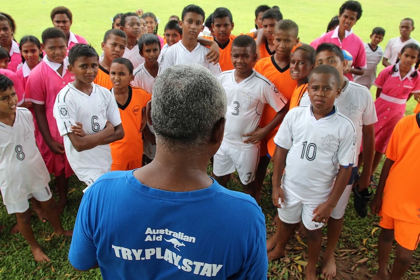 A teacher addresses a large group of students. His back is to the camera. His t-shirt says Australian Aid. Try. Play. Stay.