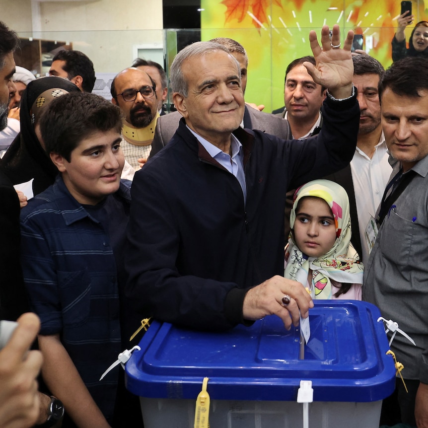  A smiling man in a suit casts a ballot at a crowded polling station.