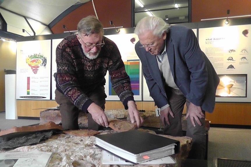 Dr Ritchie and Sir David bend over a fish fossil stone slab in a building with a lit up information display behind them.