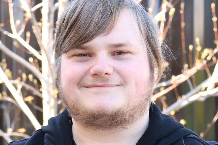 A head and shoulders shot of young Tasmanian resident Tyler Bakes, who is smiling. There is a tree in the background.