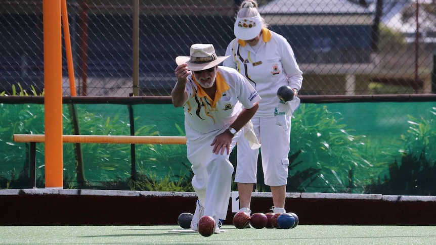 A man in white competition uniform rolls a lawn bowl towards the camera.