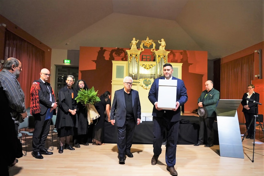 Two men walk towards the camera, one holding a large white box containing ancestral remains