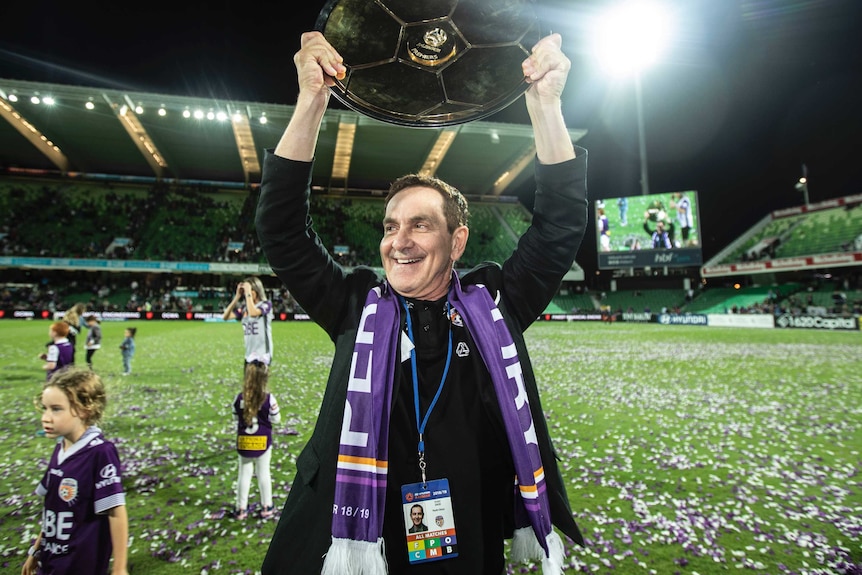 Perth Glory owner Tony Sage holds the A-League Premiers Plate aloft with a scarf around his neck as he walks on a soccer pitch.