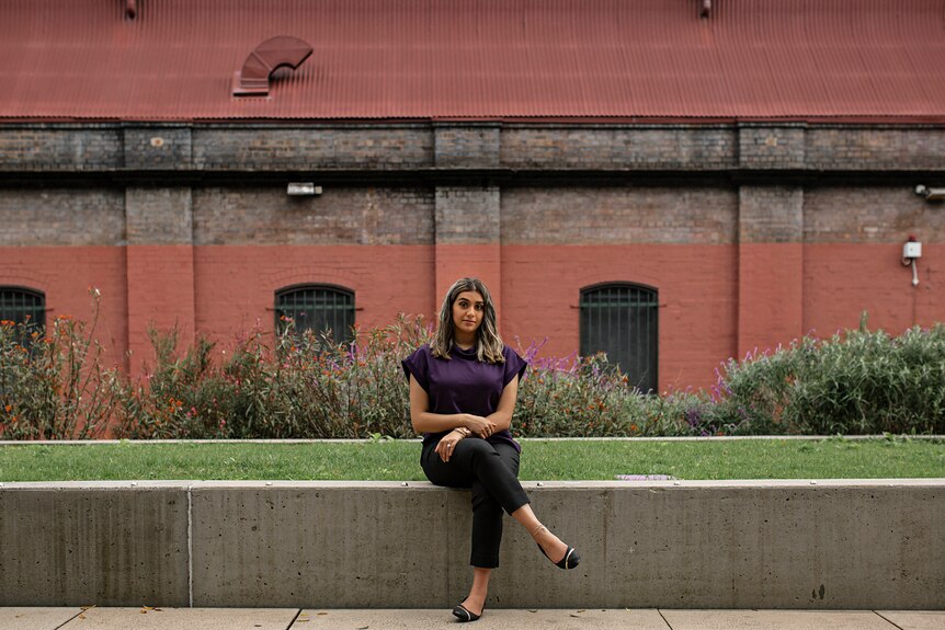 Young Muslim woman Sabreen Hussain sitting cross-legged, with garden and rust-coloured building in background.