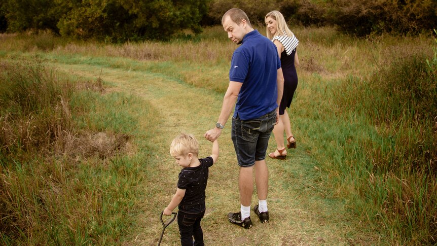 A man holds the hand of a small boy who is hauling a toy cart as they and a woman walk along a grassed path between scrub.