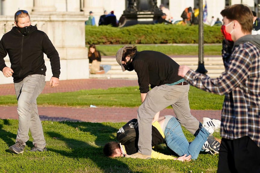 A man lays face down on the grass as another man pushes his back down.