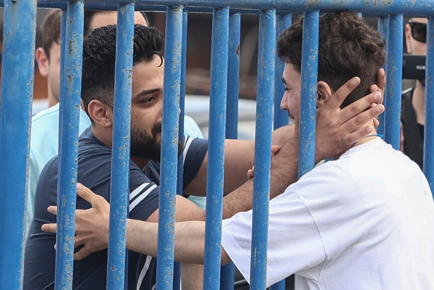a man in a navy blue shirt puts his hands through the gaps in some blue metal bars, to hold the face of a man with a white shirt