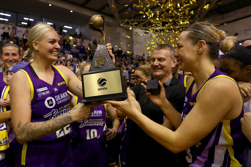 Cayla George and Tess Madgen hold the WNBL trophy and smile at each other.