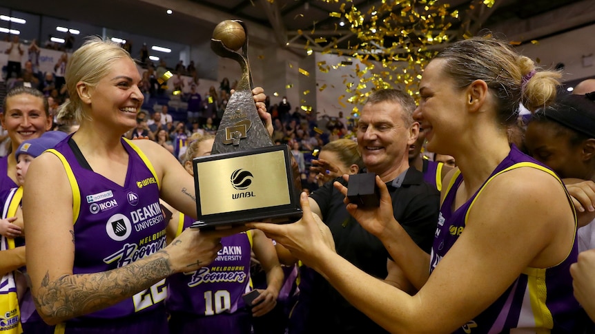Cayla George and Tess Madgen hold the WNBL trophy and smile at each other.