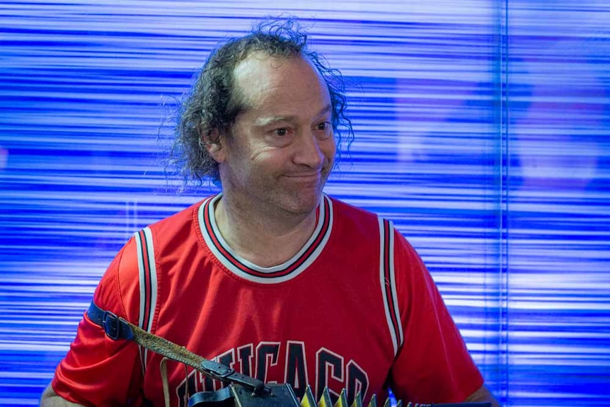 A man in a red basketball shirt plays accordion in a busy city tunnel against a blue background