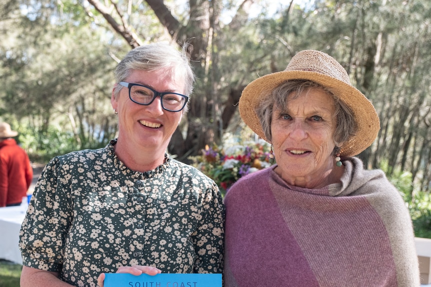Two smiling women, both grey haired, one wears glasses, green printed top, other wears hat, multi-coloured poncho, trees behind.