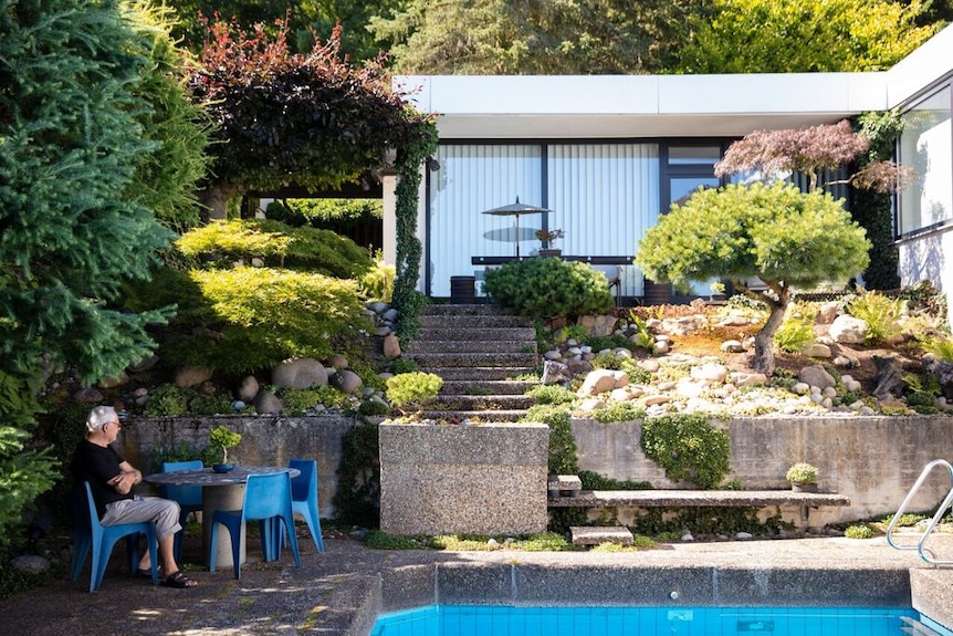 A man sits at an outdoor table surrounded by garden and a swimming pool.