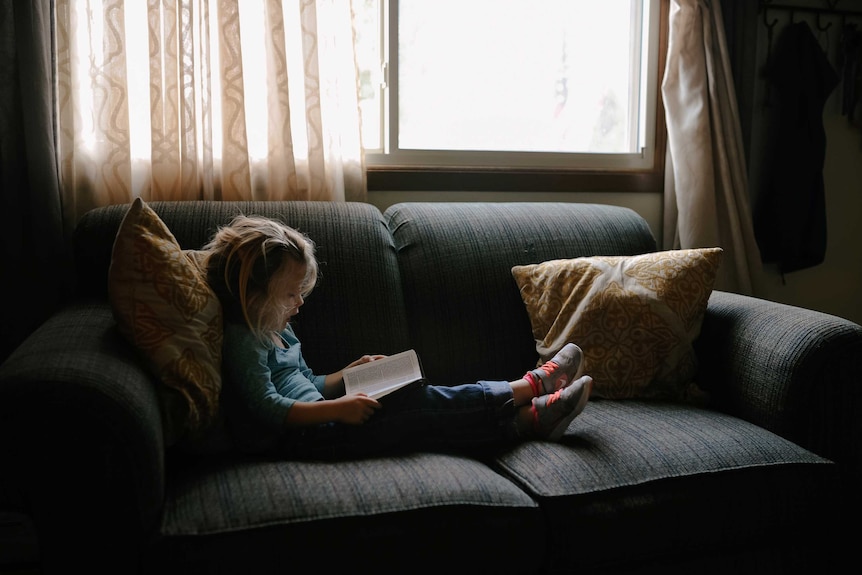 A little girl sits on a couch with her feet up, reading a book