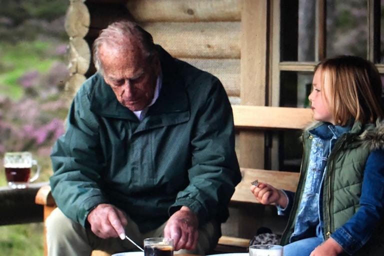 A young girl and an older man sit outside a log cabin, warmly dressed.