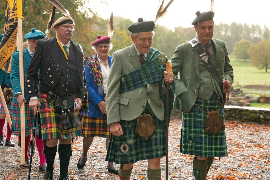 A group of people in traditional Scottish attire of kilts, coats and hats walk along a path.