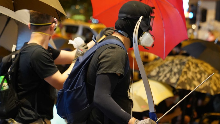 Two men clad in black are seen behind umbrellas as one carries a bow and arrow.