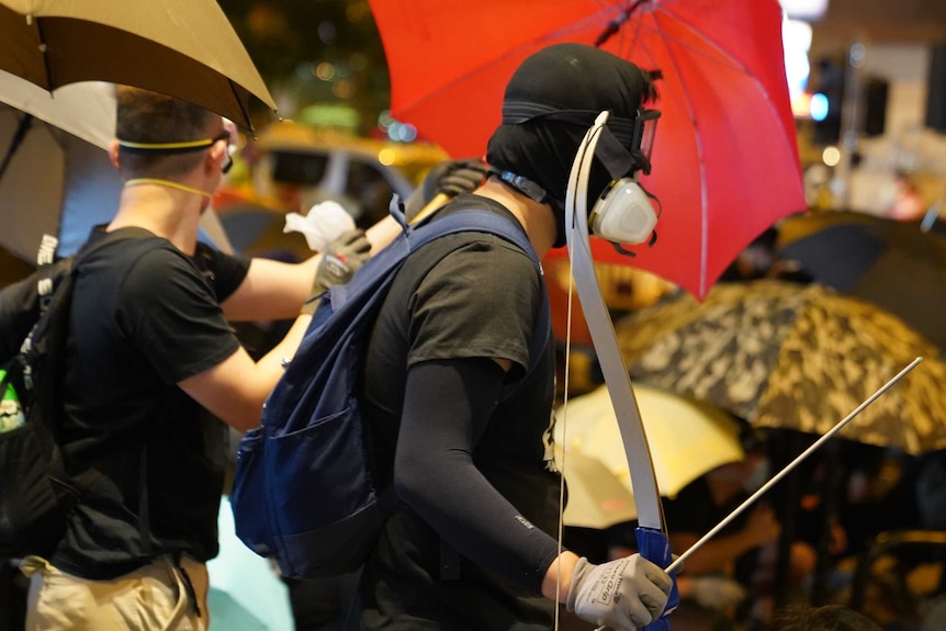 Two men clad in black are seen behind umbrellas as one carries a bow and arrow.