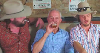 Turnbull drinks a beer while sitting on a couch in between two male journalists wearing Akubra hats