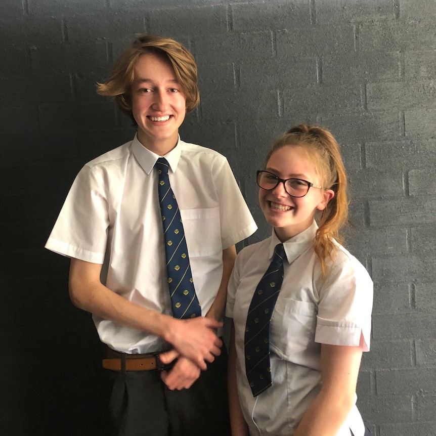 Two high school students, a boy on the left and a girl on the right, stand side by side in front of a brick wall.