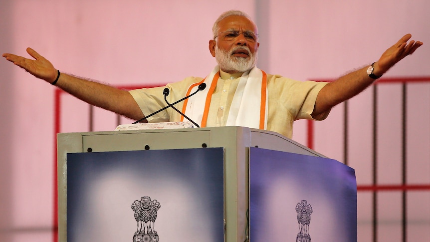 Narendra Modi gestures with arms wide and palms up as he speaks into two mics at a lectern.