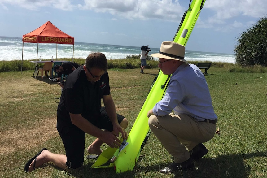 Two men crouching near a fluoro yellow flotation device, on grass on a beach foreshore.