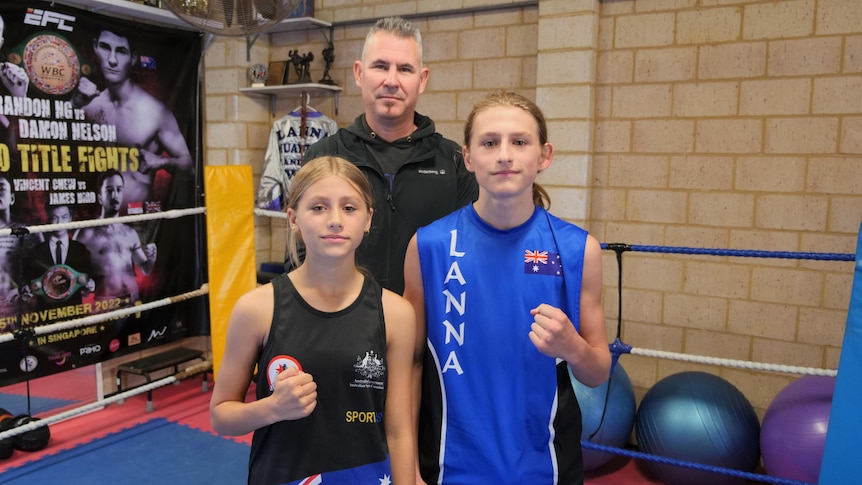 A young girl and boy stand in front of a man inside a boxing ring.
