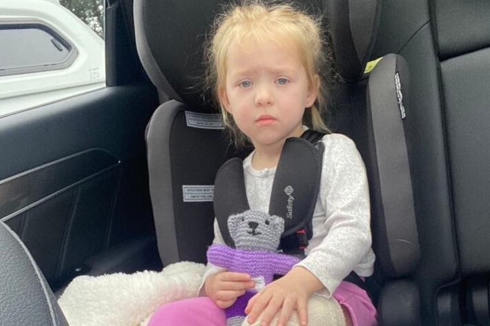 A little girl with a cast on her leg sits in a car, looking glum.
