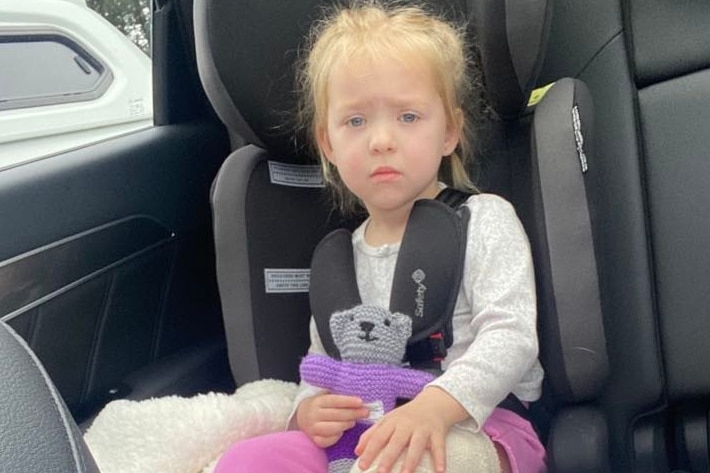 A little girl with a cast on her leg sits in a car, looking glum.