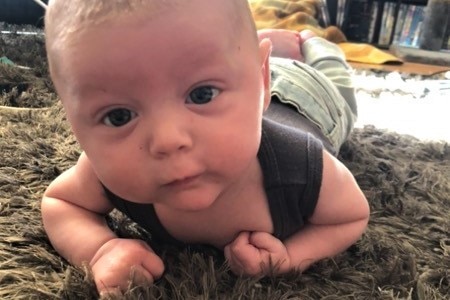 A baby lies on the carpet propping himself up on his elbows and looking at the camera