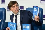 Man in navy suit holds up multiple copies of a Russian textbook at a press conference